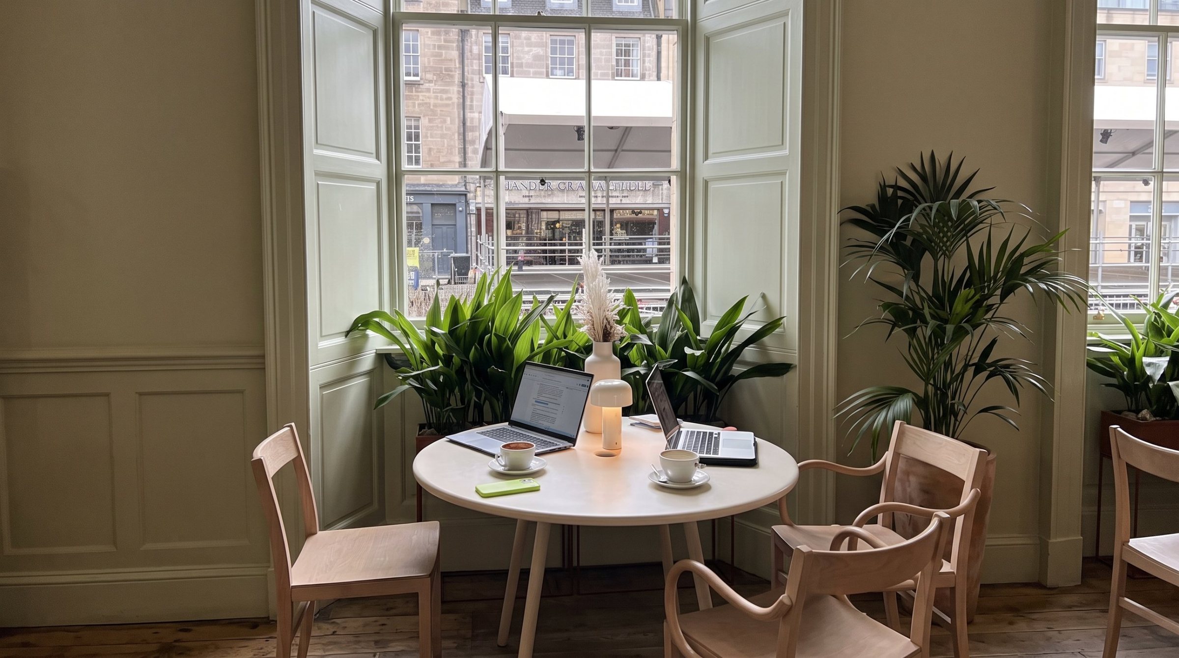 A sunlit café table by a tall window, with two laptops, coffee cups and plants